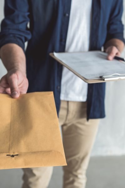 This is for you. Close-up of envelope in hand of diligent professional courier. He is standing with package and personal contract against gray wall background. Selective focus